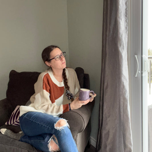 Image shows a woman sitting in a chair wearing a sweater and ripped jeans, holding a cup of tea and looking out a window 